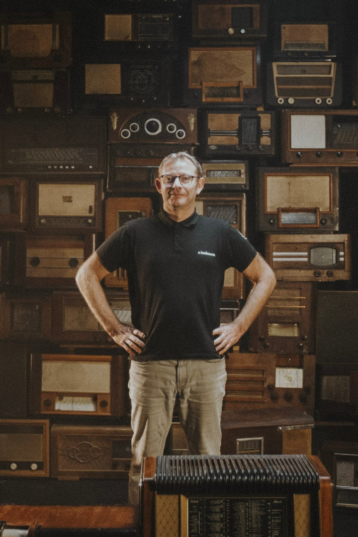 Sylvain, technicien chez A.bsolument, se tient debout devant un mur composé de radios vintage modernisées, illustrant le savoir-faire artisanal et la rencontre entre design rétro et technologie moderne.